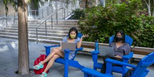 Two students engage in online classes while sitting outside wearing masks.