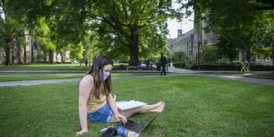 Masked girl sits on grass working on her laptop outside.