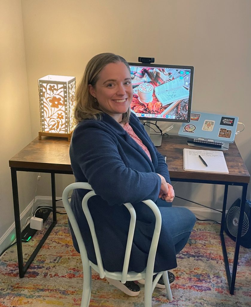 Heather Hans sitting at her desk at her home office. There is a multicolored rug, a light blue chair, a wood and metal desk, a white lamp, and a large monitor.